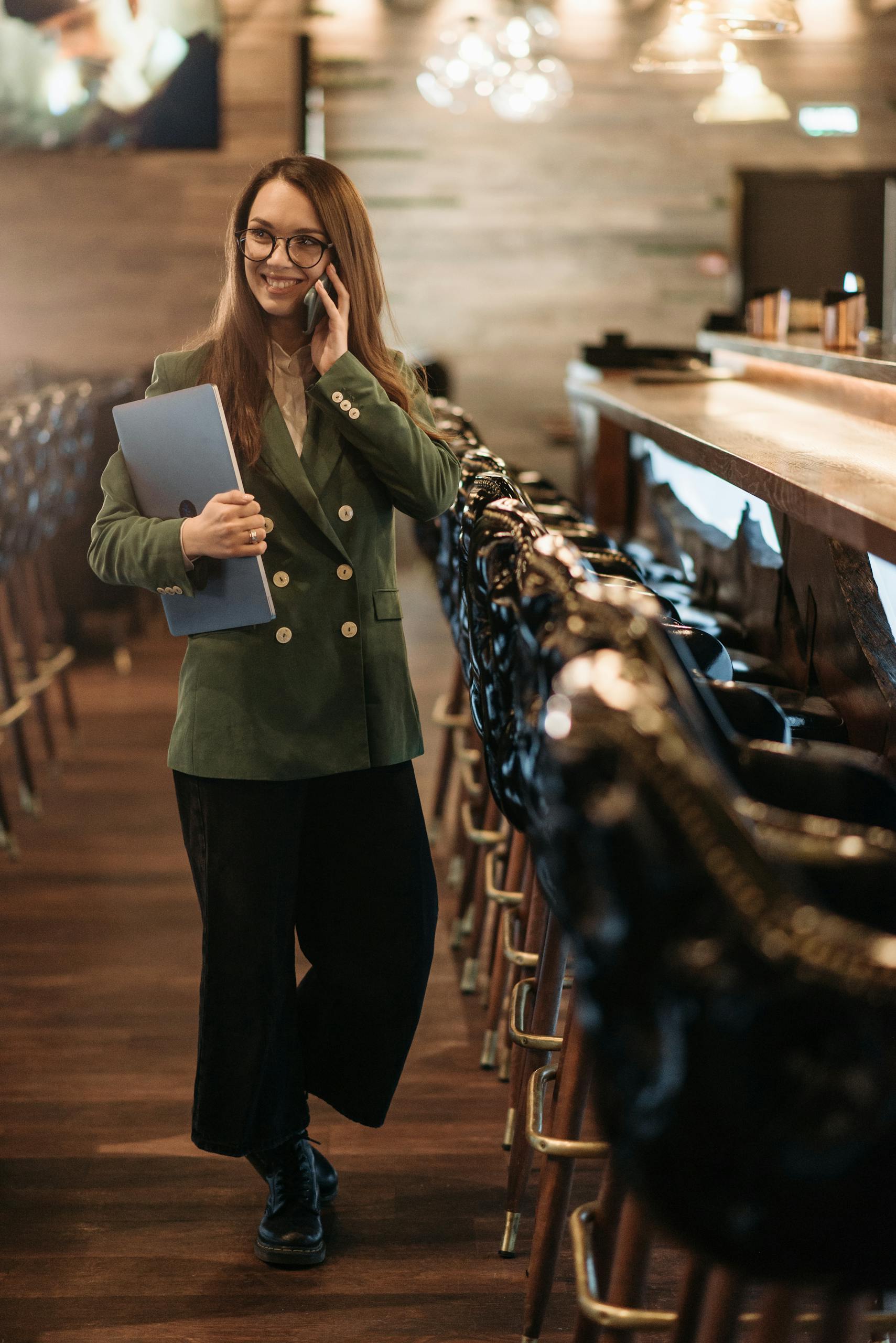 Confident businesswoman in a bar holding a laptop and talking on the phone, smiling.