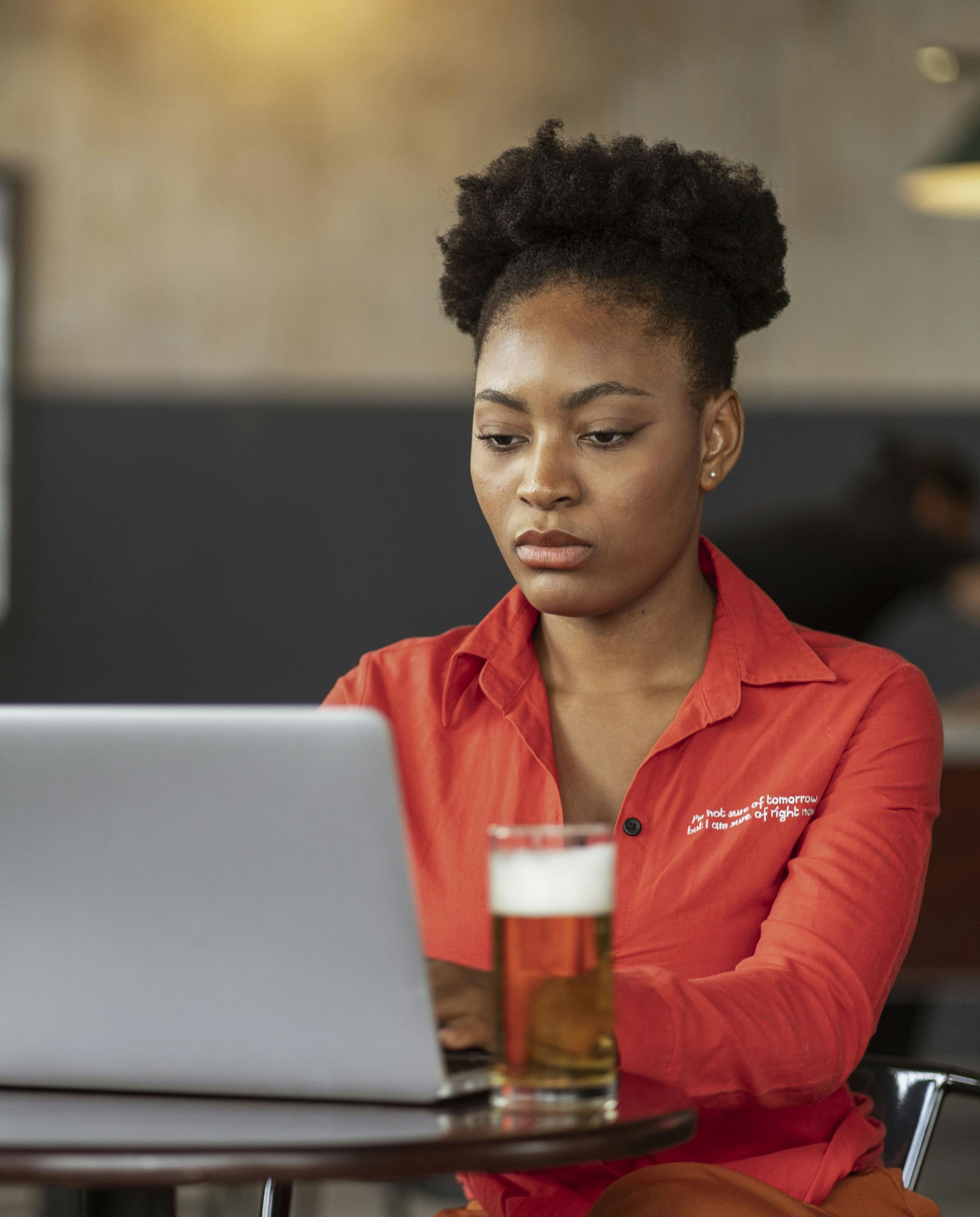Focused woman in cafe using laptop and enjoying a beer, embodies modern digital lifestyle.